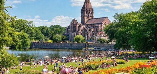 A vibrant summer scene in Magdeburg, Germany, showcasing the iconic Magdeburg Cathedral beside the Elbe River, surrounded by lush green parks filled with cheerful festival-goers enjoying picnics, music, and theater performances under the warm sun. High-definition, depicting colorful flowers, and lively activities that capture the essence of a bustling summer city.