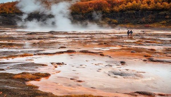 Create a high-definition image of Hell's Gate Geothermal Reserve in New Zealand during autumn. The scene should capture the vibrant geothermal features like steaming fumaroles and bubbling mud pools, set against a backdrop of warm-hued foliage as the leaves change color. Include a serene atmosphere with a few visitors exploring the landscape, embodying a mix of rich Maori culture and natural beauty. The image should evoke a sense of tranquility and adventure in this enchanting environment.