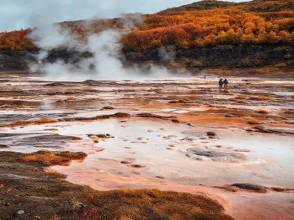 Create a high-definition image of Hell's Gate Geothermal Reserve in New Zealand during autumn. The scene should capture the vibrant geothermal features like steaming fumaroles and bubbling mud pools, set against a backdrop of warm-hued foliage as the leaves change color. Include a serene atmosphere with a few visitors exploring the landscape, embodying a mix of rich Maori culture and natural beauty. The image should evoke a sense of tranquility and adventure in this enchanting environment.