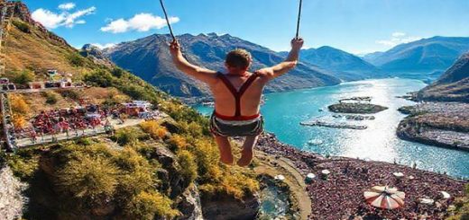"Create a high definition image of AJ Hackett Bungy in Queenstown, New Zealand, showcasing the thrilling bungy jump against a breathtaking seasonal backdrop. Depict summer with vibrant colors, the lush landscape, and clear blue skies, filled with excited jumpers and bustling crowds. Capture the sparkling waters of Lake Wakatipu reflecting the sun, with hints of nearby music festivals that add liveliness to the scene."