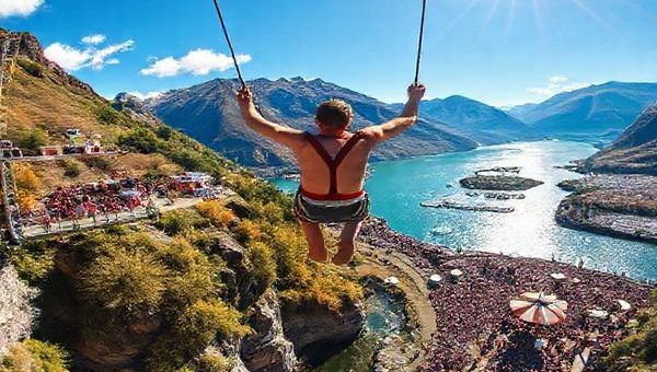 "Create a high definition image of AJ Hackett Bungy in Queenstown, New Zealand, showcasing the thrilling bungy jump against a breathtaking seasonal backdrop. Depict summer with vibrant colors, the lush landscape, and clear blue skies, filled with excited jumpers and bustling crowds. Capture the sparkling waters of Lake Wakatipu reflecting the sun, with hints of nearby music festivals that add liveliness to the scene."