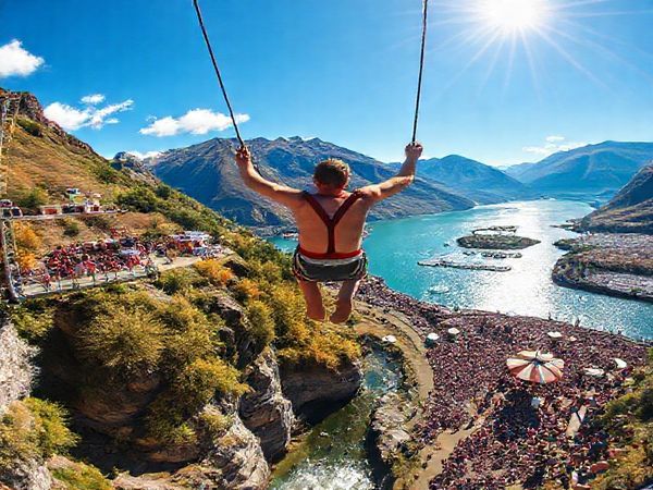 "Create a high definition image of AJ Hackett Bungy in Queenstown, New Zealand, showcasing the thrilling bungy jump against a breathtaking seasonal backdrop. Depict summer with vibrant colors, the lush landscape, and clear blue skies, filled with excited jumpers and bustling crowds. Capture the sparkling waters of Lake Wakatipu reflecting the sun, with hints of nearby music festivals that add liveliness to the scene."