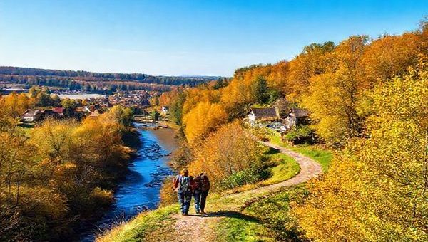 A breathtaking high-definition view of Landkreis Oberspreewald-Lausitz, showcasing its picturesque landscapes during autumn. Vibrant trees in shades of gold and amber line a serene stream, while quaint villages with charming old buildings dot the scenery. A couple enjoys a peaceful hike along a winding path that leads through lush woods, with the fresh, crisp air surrounding them. In the background, a clear blue sky hints at the unpredictable yet invigorating nature of this idyllic German region.
