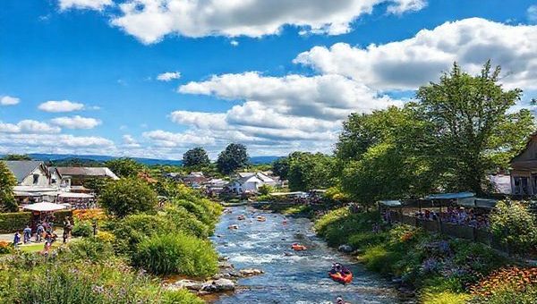 A vibrant summer scene in Kaiapoi, New Zealand, showcasing the picturesque river flowing through the town surrounded by lush greenery and colorful flowers. Bright blue skies overhead filled with fluffy white clouds, with people kayaking on the river and picnicking in the beautiful Kaiapoi Domain park. Include local art exhibitions and community events bustling with cheerful visitors, all captured in stunning high definition.
