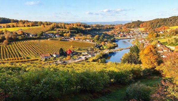 A picturesque landscape of the Rhein-Neckar-Kreis region in southwestern Germany, showcasing sprawling vineyards under warm summer sunshine, quaint villages with vibrant festivals, and serene hiking trails surrounded by colorful autumn foliage. Incorporate elements like the Neckar River for picnicking and traditional architectural marvels to reflect historical charm. Create this image in high definition, capturing the essence of each season's beauty.