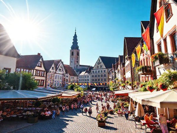 Create a high-definition image of the charming city of Fürth in Bavaria, Germany, showcasing the vibrant summer atmosphere. Picture bustling plazas filled with outdoor cafes serving traditional Bavarian treats, radiant sunshine illuminating the cobblestone streets, and the iconic architectural landmarks of the city. Include details like locals enjoying outdoor activities by the Pegnitz River and colorful banners from the Michaeliskirchweih fair fluttering in the breeze, conveying a lively, cheerful scene.