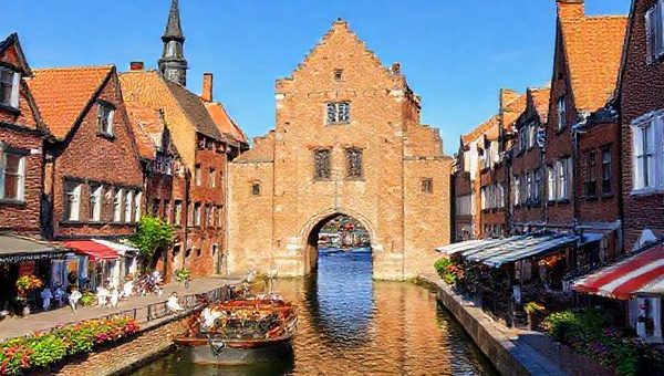 A picturesque view of Lübeck, showcasing its stunning brick Gothic architecture and narrow cobbled streets, bathed in the warm glow of summer sunlight. The scene features the iconic Holsten Gate against a clear blue sky, with tourists enjoying a boat tour along the tranquil canals and vibrant flowers blooming along the banks. The atmosphere is lively yet inviting, with quaint bakeries visible along the streets. Generate this image in high definition.