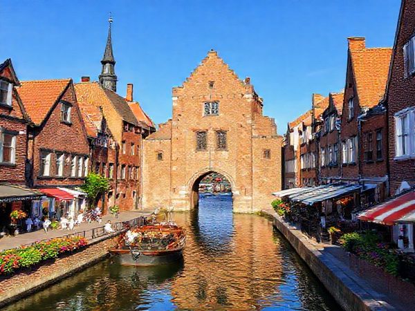 A picturesque view of Lübeck, showcasing its stunning brick Gothic architecture and narrow cobbled streets, bathed in the warm glow of summer sunlight. The scene features the iconic Holsten Gate against a clear blue sky, with tourists enjoying a boat tour along the tranquil canals and vibrant flowers blooming along the banks. The atmosphere is lively yet inviting, with quaint bakeries visible along the streets. Generate this image in high definition.