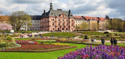 A picturesque scene of Darmstadt in spring, showcasing blooming flowers and lush greenery, with the iconic art nouveau buildings of Mathildenhöhe in the background. Include people leisurely strolling through the vibrant parks, enjoying cultural exhibitions, and a hint of a cloudy sky giving way to sunshine. Capture the essence of renewal and outdoor exploration, ensuring the image is in high definition.