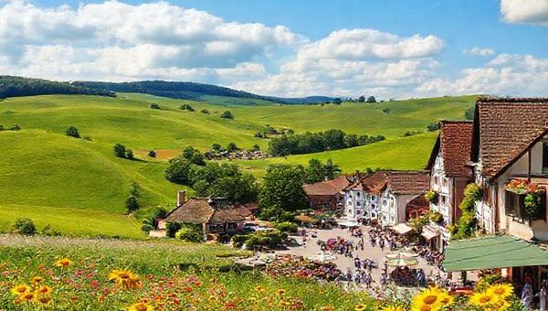 A high-definition image of the picturesque Landkreis Sömmerda in Thuringia, Germany, showcasing a vibrant summer scene with lush green rolling hills, fields of wildflowers and sunflowers dancing in the breeze, and quaint village life. Include a historical town square bustling with a local festival, colorful stalls offering traditional food, and people enjoying music and dance under a clear blue sky filled with soft, fluffy clouds. Add a touch of magic by capturing the warmth of the sun and a sense of pastoral tranquility.
