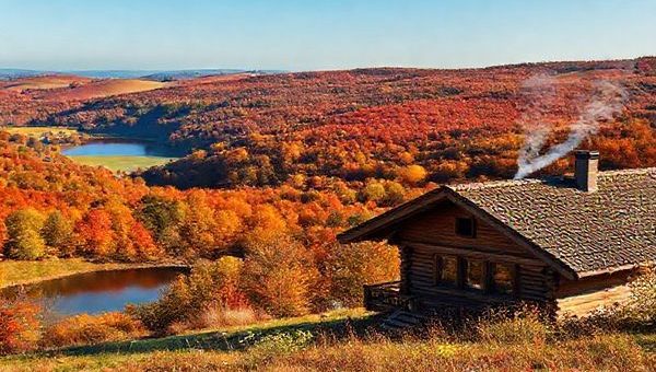 A serene landscape of Landkreis Uckermark in autumn, showcasing a picturesque scene of rolling hills draped in vibrant shades of red, orange, and gold foliage. Include a tranquil lake reflecting the colorful trees, with a few scattered wildflowers blooming nearby. Capture a cozy wooden cabin with smoke gently rising from the chimney, surrounded by nature and a clear blue sky. High definition.