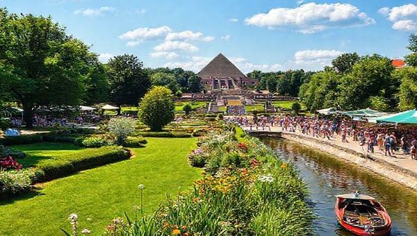 A breathtaking high-definition image of Cottbus, Germany, showcasing the city's vibrant charm in summer: lush gardens of Branitz Park with the Pückler Pyramids in the background, lively people mingling during a cultural festival, and the serene Spreewald waterways inviting visitors for boat rides. The scene is filled with bright sunshine, colorful flowers, and festive decorations, encapsulating the essence of a bustling summer day in this hidden gem of Europe.