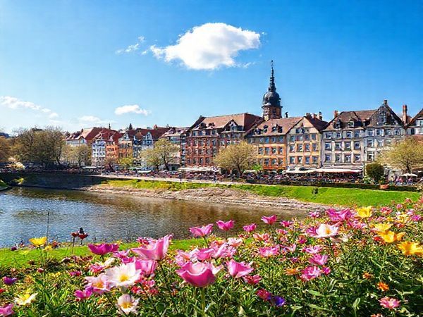 A high-definition image of Saarbrücken in spring, showcasing vibrant blooming flowers in lush parks, with the scenic Saar River flowing nearby. Capture the lively atmosphere with locals enjoying outdoor cafés and cyclists on paths, under a bright blue sky with a few fluffy clouds. Feature charming historical buildings in the background, all bathed in soft, warm sunlight, symbolizing rejuvenation and the beauty of this enchanting German city.