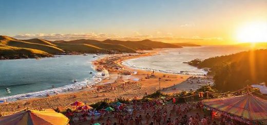 A breathtaking high-definition scene of the Ōpōtiki District, New Zealand, showcasing the vibrant beauty of its summer landscape. Picture golden beaches bustling with sunbathers and surfers, surrounded by lush rolling hills and distinctive native forests. Include a lively cultural festival in the foreground, with colorful Maori decorations and people dancing. The sun casts a warm glow on the horizon, enhancing the overall festive atmosphere of this serene coastal gem.