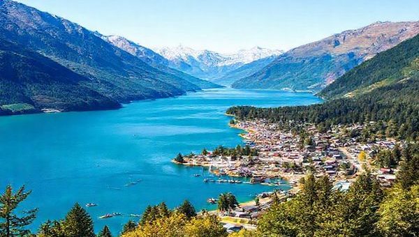 A breathtaking high-definition landscape of Queenstown-Lakes District in summer, showcasing the vibrant blue waters of Lake Wakatipu surrounded by rugged mountains and lush green forests. Include adventure seekers kayaking and paddleboarding on the lake. The town of Queenstown buzzes with energy, featuring outdoor festivals and colorful market stalls under a clear, sunny sky. Capture the essence of summer vibrancy and natural beauty in this paradise setting.