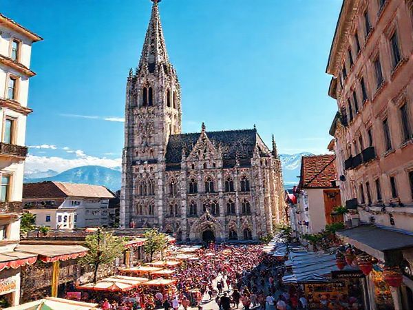 A majestic view of Ulm Minster soaring under a brilliant blue summer sky, with the intricate stone carvings of its Gothic façade illuminated by the sun. Lively street festivals are taking place below, filled with colorful decorations and bustling crowds. In the background, the distant Alps shimmer in the clear weather, creating a breathtaking vista. Render in high definition.