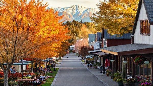 A picturesque scene of Rangiora, New Zealand, showcasing the vibrant beauty of all four seasons: summer with sunlit parks filled with picnicking families, fall with amber and gold foliage, winter featuring cozy cafes with steaming cups of coffee, and spring bursting with blooming flowers. Include the Southern Alps in the distance, creating a dramatic backdrop. The image should capture the warm community spirit and scenic charm of this quaint town in high definition.