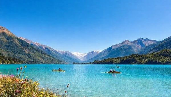 A breathtaking high-definition landscape of Lake Clearwater, New Zealand, showcasing the serenity of summer with vibrant azure waters and surrounding lush greenery, framed by majestic mountains under a clear blue sky. Include water sports enthusiasts kayaking and windsurfing, alongside blooming wildflowers and active wildlife like black stilts. The scene should evoke a sense of adventure and tranquility, capturing the essence of this hidden gem in Canterbury.