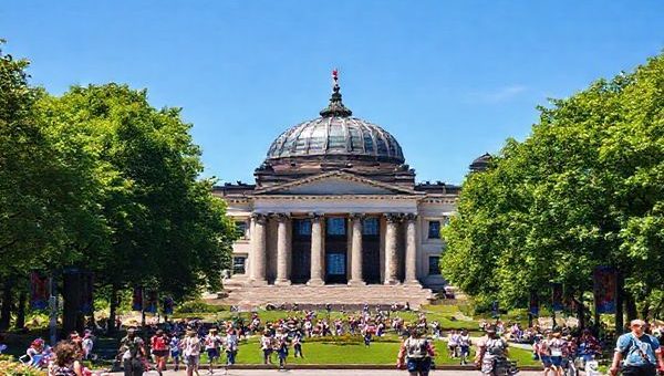 A high definition image of the Reichstag Building in summer, surrounded by vibrant green foliage of Tiergarten park and busy tourists enjoying the sunny day. The iconic glass dome glistens under the bright blue sky, with people walking around, capturing the lively atmosphere of Berlin in summer. Include elements like picnic blankets, bicycles, and blooming flowers to emphasize the energetic vibe of the season.