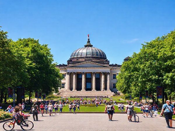 A high definition image of the Reichstag Building in summer, surrounded by vibrant green foliage of Tiergarten park and busy tourists enjoying the sunny day. The iconic glass dome glistens under the bright blue sky, with people walking around, capturing the lively atmosphere of Berlin in summer. Include elements like picnic blankets, bicycles, and blooming flowers to emphasize the energetic vibe of the season.