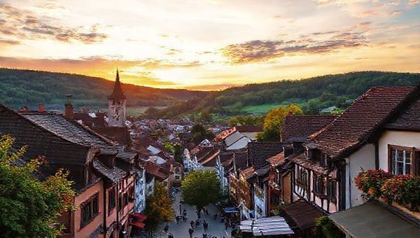 A picturesque view of Arnsberg, Germany, showcasing its charming medieval architecture and lush green hills under a vibrant sunset sky, with quaint streets bustling with people enjoying the warm ambiance of early evening. Include seasonal elements like blooming flowers in spring, colorful autumn leaves, and a cozy outdoor café scene. Ensure the image is in high definition.