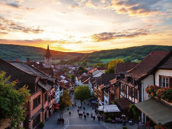 A picturesque view of Arnsberg, Germany, showcasing its charming medieval architecture and lush green hills under a vibrant sunset sky, with quaint streets bustling with people enjoying the warm ambiance of early evening. Include seasonal elements like blooming flowers in spring, colorful autumn leaves, and a cozy outdoor café scene. Ensure the image is in high definition.