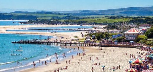 A high-definition image of the picturesque Timaru District in New Zealand, showcasing a vibrant summer scene at Caroline Bay. Capture families enjoying the beach with sunbathers, children playing on the sand, and water sports like paddleboarding and sailing. In the background, lush green landscapes and the gentle waves of the sea extend towards hazy mountains under a bright blue sky. Include elements of the lively Caroline Bay Carnival, with colorful rides and joyful crowds, reflecting the playful spirit of summer.