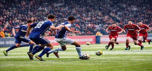 A dramatic football scene capturing the clash between Ross County and Airdrie on a snowy December day in Dingwall, Scotland. Visually portray Ross County's resilient defense, with players in navy jerseys forming a compact line, contrasted against Airdrie's dynamic forward attack in bold red kits rushing towards the goal. Include a vibrant crowd in the background, embodying the electric atmosphere, while snowflakes gently fall around the stadium. Ensure the image is in high definition.