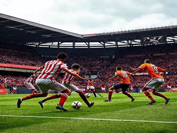 **High definition image of a dramatic football scene depicting a tense League Two match between Fleetwood Town and Salford City. The stadium is packed with passionate fans waving team colors, under a cloudy December sky. On the pitch, two teams are locked in a fierce battle; Fleetwood players, wearing their traditional red and white stripes, are focusing intently on defense, while Salford City players, donned in vibrant orange, are poised for a dynamic attack. Capture the intensity of the moment - a midfielder making a tactical interception, a forward preparing to sprint for goal, with the scoreboards reflecting the high stakes of this critical match.**