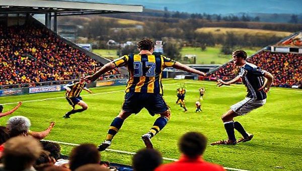 A vibrant high-definition scene depicting a fierce Scottish League One football match between Alloa Athletic and Peterhead. Capture the moment of intense action on the pitch, highlighting Alloa's dynamic midfield maestro orchestrating a fluid attack with creative passing, while Peterhead's robust captain commands the defensive line, ready for a counterattack. The stadium is packed with passionate fans in colorful jerseys, exuding palpable tension and excitement. The backdrop features the picturesque Scottish landscape, adding to the lively atmosphere of this clash of footballing philosophies.