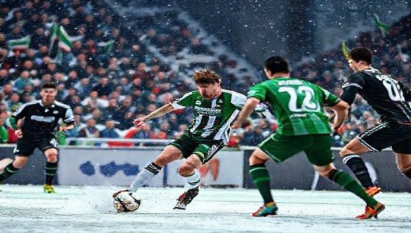 Create a high-definition image of a dramatic soccer match between Breidablik and Shamrock Rovers. Capture the moment where Breidablik's captain, a determined Icelandic player, makes a swift counter-attack on a snowy pitch, with the Shamrock Rovers players positioned defensively in an urban Dublin stadium atmosphere. The crowd is vibrant, immersed in anticipation, with flags waving and a sense of rivalry in the air. Highlight the contrasting styles: Breidablik's gritty resilience against Shamrock Rovers' strategic sophistication, showcasing the intensity and dynamic energy of the Europa Conference League clash.