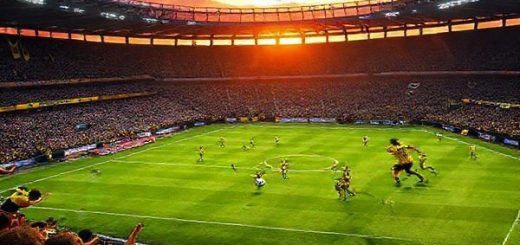 A high-definition image depicting an electrifying football match scene between Borussia Dortmund and Bodo/Glimt in a packed stadium. The atmosphere is charged with energy, showcasing Dortmund's dynamic young players sprinting with fervor against Bodo/Glimt's organized and disciplined defense. Capture the contrasting styles: Dortmund's fiery, aggressive play versus Bodo/Glimt's calculated strategy. Include fans in vibrant team colors, banners waving, and a dramatic sunset casting vivid colors over the pitch, highlighting the tension of this Champions League showdown.