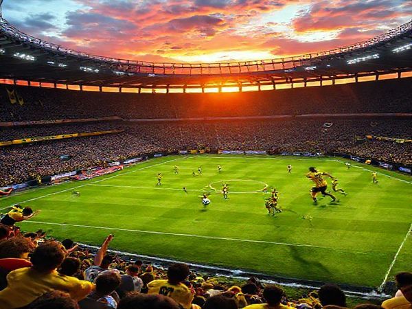 A high-definition image depicting an electrifying football match scene between Borussia Dortmund and Bodo/Glimt in a packed stadium. The atmosphere is charged with energy, showcasing Dortmund's dynamic young players sprinting with fervor against Bodo/Glimt's organized and disciplined defense. Capture the contrasting styles: Dortmund's fiery, aggressive play versus Bodo/Glimt's calculated strategy. Include fans in vibrant team colors, banners waving, and a dramatic sunset casting vivid colors over the pitch, highlighting the tension of this Champions League showdown.