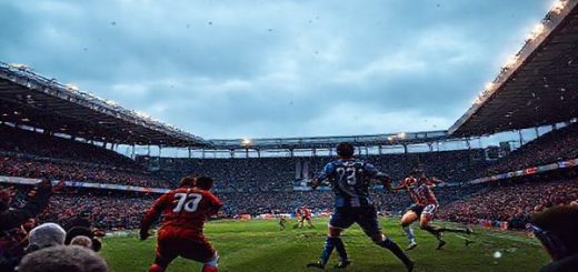 "Create a high-definition image of an intense Scottish League Two football match between Edinburgh City and Dumbarton. Capture the vibrant atmosphere of December, with passionate fans cheering in a packed stadium, players in dynamic action representing Edinburgh City's high-energy press and Dumbarton's disciplined defense. Include details like the players' jerseys—Edinburgh in their spirited colors and Dumbarton in their classic hues— under a moody, wintry sky. Emphasize the tension of the match, showcasing the clash of styles and the determination of both teams."