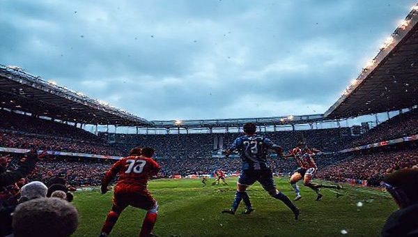 "Create a high-definition image of an intense Scottish League Two football match between Edinburgh City and Dumbarton. Capture the vibrant atmosphere of December, with passionate fans cheering in a packed stadium, players in dynamic action representing Edinburgh City's high-energy press and Dumbarton's disciplined defense. Include details like the players' jerseys—Edinburgh in their spirited colors and Dumbarton in their classic hues— under a moody, wintry sky. Emphasize the tension of the match, showcasing the clash of styles and the determination of both teams."