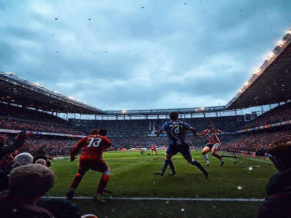 "Create a high-definition image of an intense Scottish League Two football match between Edinburgh City and Dumbarton. Capture the vibrant atmosphere of December, with passionate fans cheering in a packed stadium, players in dynamic action representing Edinburgh City's high-energy press and Dumbarton's disciplined defense. Include details like the players' jerseys—Edinburgh in their spirited colors and Dumbarton in their classic hues— under a moody, wintry sky. Emphasize the tension of the match, showcasing the clash of styles and the determination of both teams."