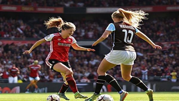 Create a high-definition image capturing a dynamic soccer match scene between Manchester United Women and West Ham Women. The field is alive with energy as players are in motion—Manchester United’s Ella Toone orchestrates a swift attack while West Ham’s Dagny Brynjarsdottir stands resolute in defense. The vibrant stadium backdrop is filled with enthusiastic fans, banners waving, and a sense of palpable excitement in the air, illustrating the clash of ambition and tactical prowess on the pitch.