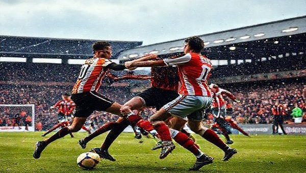 A dramatic scene unfolds at Brisbane Road under chilly December skies, showcasing a fiercely contested football match between Leyton Orient and Luton Town. Players from both teams clash on the field, with Leyton Orient’s resilient defenders standing firm against Luton's dynamic attackers. In the background, a stadium filled with passionate fans, representing the vibrant colors of both teams, adds to the electric atmosphere. The tension is palpable, capturing the essence of this League One showdown amid the excitement of World Cup qualifiers. Render this image in high definition.