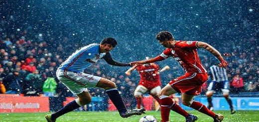 **Prompt:** A dramatic football scene capturing the intense atmosphere of a League One match between Cardiff City and Huddersfield Town on a cold December day in 2025. The pitch is alive with the energy of passionate fans in vibrant colors, a tense showdown between players showcasing Cardiff's disciplined strategy against Huddersfield's unpredictable flair. Snowflakes gently fall as the teams engage in a tactical battle akin to chess on grass. Make the image in high definition.