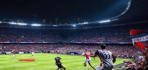 A high-definition image of a thrilling Champions League football match between Qarabag FK and Ajax at night in Baku. The stadium is lit with vibrant colors, filled with excited fans waving flags, while players embody the clash of styles: Qarabag's disciplined defense, led by Maksim Medvedev, contrasts with Ajax's fluid attack, featuring Brian Brobbey. The atmosphere is electric, showcasing strategic intensity and the spirit of competition, set against a dramatic city skyline.
