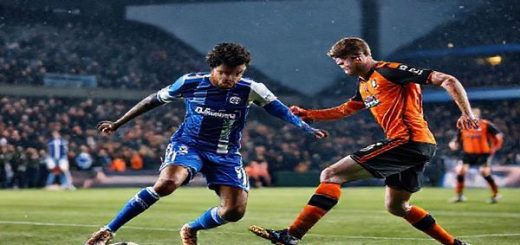High definition image of a dramatic football match scene set on a cold December night, showcasing Olympique Lyonnais in their signature blue and white kits, elegantly orchestrating a play against Go Ahead Eagles in their vibrant orange and black uniforms. The stadium is filled with passionate fans cheering, with a backdrop of city lights, a competitive atmosphere, and dynamic player movement captured mid-action. Include a close-up of key players: Lyon’s Houssem Aouar controlling the ball and Go Ahead Eagles’ Oliver Edvardsen preparing for a counter-attack, highlighting the clash of contrasting football philosophies.
