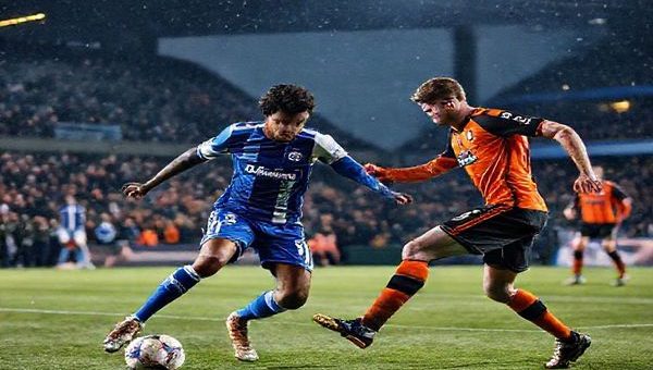 High definition image of a dramatic football match scene set on a cold December night, showcasing Olympique Lyonnais in their signature blue and white kits, elegantly orchestrating a play against Go Ahead Eagles in their vibrant orange and black uniforms. The stadium is filled with passionate fans cheering, with a backdrop of city lights, a competitive atmosphere, and dynamic player movement captured mid-action. Include a close-up of key players: Lyon’s Houssem Aouar controlling the ball and Go Ahead Eagles’ Oliver Edvardsen preparing for a counter-attack, highlighting the clash of contrasting football philosophies.