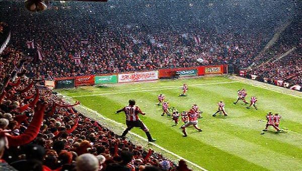 A high-definition image depicting a tense moment during the League Two match between Swindon Town and Cheltenham Town in December 2025. Capture a crowded, vibrant stadium filled with passionate fans waving team colors, as players from both teams engage in a fierce midfield battle, showcasing Swindon’s tenacious defense against Cheltenham’s strategic offense. The atmosphere is electric, with snow gently falling, and festive decorations subtly incorporated, symbolizing the height of the football season.