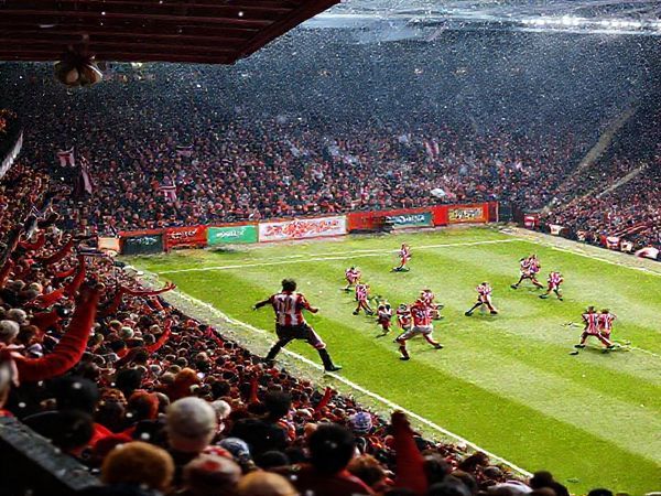 A high-definition image depicting a tense moment during the League Two match between Swindon Town and Cheltenham Town in December 2025. Capture a crowded, vibrant stadium filled with passionate fans waving team colors, as players from both teams engage in a fierce midfield battle, showcasing Swindon’s tenacious defense against Cheltenham’s strategic offense. The atmosphere is electric, with snow gently falling, and festive decorations subtly incorporated, symbolizing the height of the football season.