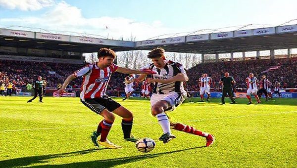 A vibrant football pitch under the bright December sky, with Bromley and Crawley players clashing in a dynamic moment of the match. The atmosphere is electric with passionate fans in the background, showcasing team colors and banners, while coaches Kevin Watson and Dan Taylor strategize from the sidelines. Capture the intense expressions of Tommy Smith and Eden Saunders as they battle for possession, surrounded by energy and determination. High definition.