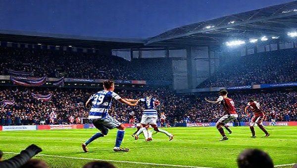 High definition image of a dramatic football match at Madejski Stadium, December 2025, featuring Reading FC in blue and white stripes clashing against Peterborough United in their iconic maroon and white kit. The stadium is buzzing with vociferous fans, waving banners, and scarves, under a crisp winter sky. Capture the essence of intense competition as players engage in a tactical battle on the pitch, with George Puscas and Jonson Clarke-Harris prominently in action. Emphasize the emotions of anticipation, resilience, and ambition in the atmosphere.