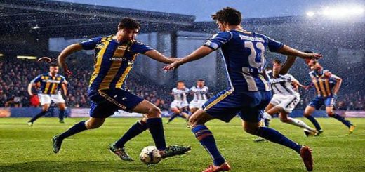 A high-definition image depicting an intense football match between Mansfield Town and Bolton Wanderers, set in a winter atmosphere under stadium lights. Capture the gritty determination of Mansfield players in their blue and amber kit, defending valiantly against Bolton's elegant, possession-focused play in white and navy. Showcase the tactical ballet between the two teams, highlighting key players like Tom Anderson and MJ Williams in action, with fans passionately cheering in the stands, and an electric, competitive vibe radiating from the scene.
