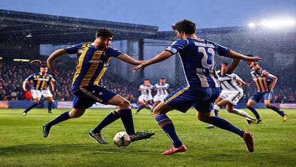 A high-definition image depicting an intense football match between Mansfield Town and Bolton Wanderers, set in a winter atmosphere under stadium lights. Capture the gritty determination of Mansfield players in their blue and amber kit, defending valiantly against Bolton's elegant, possession-focused play in white and navy. Showcase the tactical ballet between the two teams, highlighting key players like Tom Anderson and MJ Williams in action, with fans passionately cheering in the stands, and an electric, competitive vibe radiating from the scene.