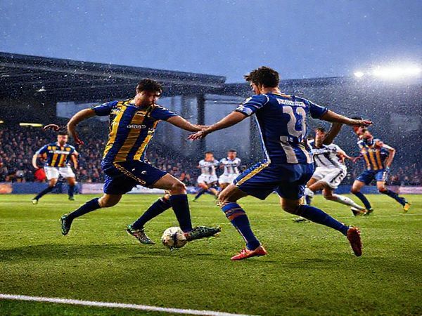 A high-definition image depicting an intense football match between Mansfield Town and Bolton Wanderers, set in a winter atmosphere under stadium lights. Capture the gritty determination of Mansfield players in their blue and amber kit, defending valiantly against Bolton's elegant, possession-focused play in white and navy. Showcase the tactical ballet between the two teams, highlighting key players like Tom Anderson and MJ Williams in action, with fans passionately cheering in the stands, and an electric, competitive vibe radiating from the scene.