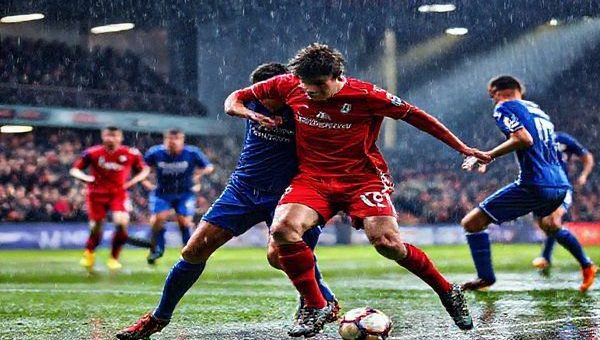 A high-definition image of a thrilling football match scene: two teams in vibrant kits, Stevenage in bright red and Cardiff in deep blue, clashing on a rain-soaked pitch under stadium lights. The atmosphere is electric, with passionate fans cheering from the stands. Focus on an intense moment where a Stevenage player tackles a Cardiff striker, showcasing the determination and skill of both teams. Capture the dynamic energy and emotion of the game, with droplets of rain reflecting the colorful lights.